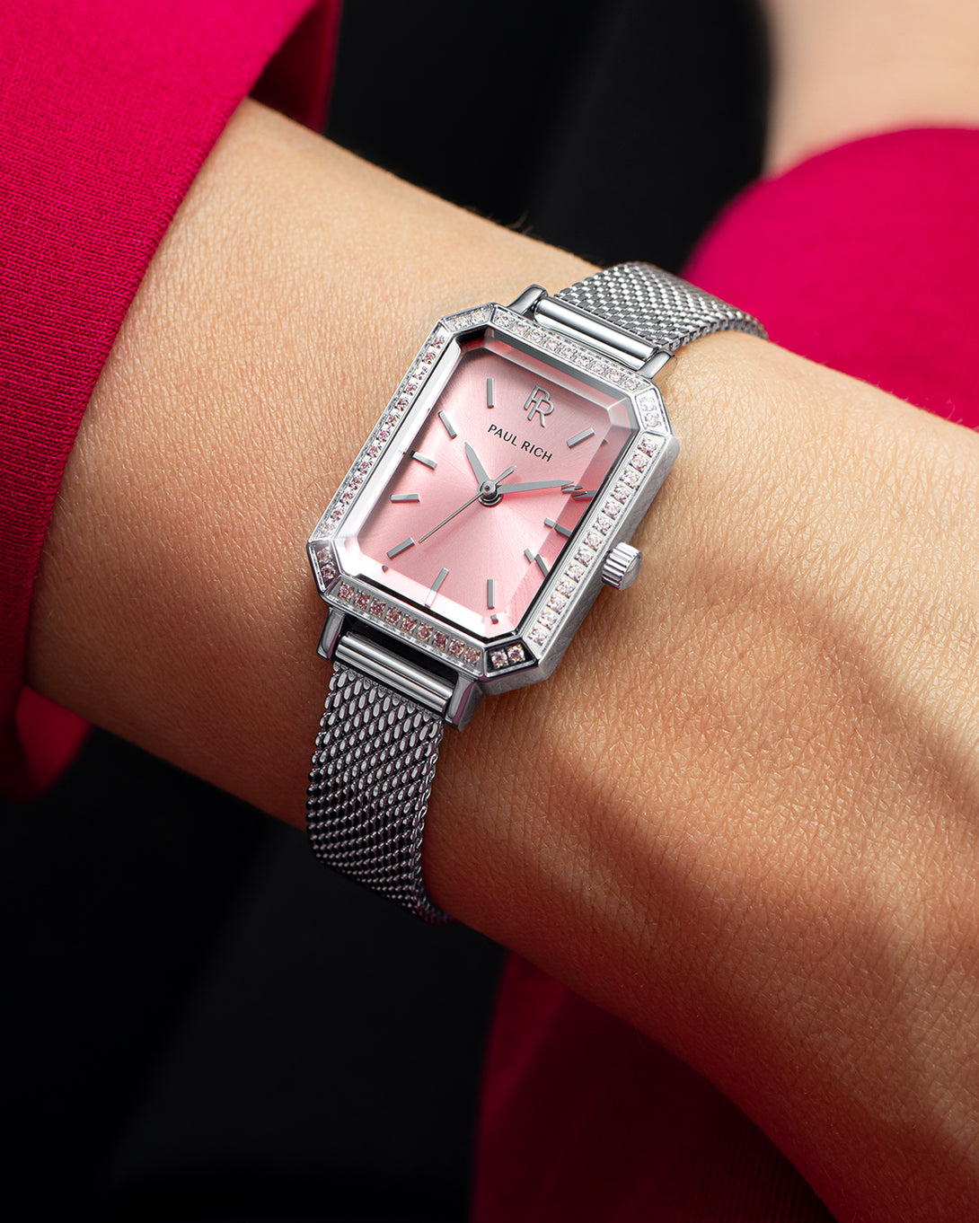 Silver-tone Paul Rich watch with pink sunray dial, pink gem-set octagonal bezel, and silver mesh strap, worn on wrist with a vibrant pink sleeve. Silver-tone Paul Rich watch with pink sunray dial, pink gem-set octagonal bezel, and silver mesh strap, worn on wrist with a vibrant pink sleeve.