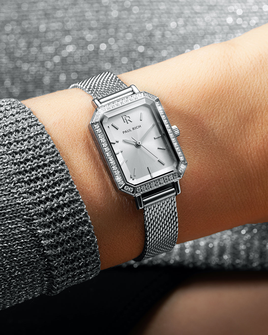 Silver-tone Paul Rich watch with silver sunray dial, gem-set octagonal bezel, and silver mesh strap, worn on wrist with a sparkling silver knit sleeve. Silver-tone Paul Rich watch with silver sunray dial, gem-set octagonal bezel, and silver mesh strap, worn on wrist with a sparkling silver knit sleeve.