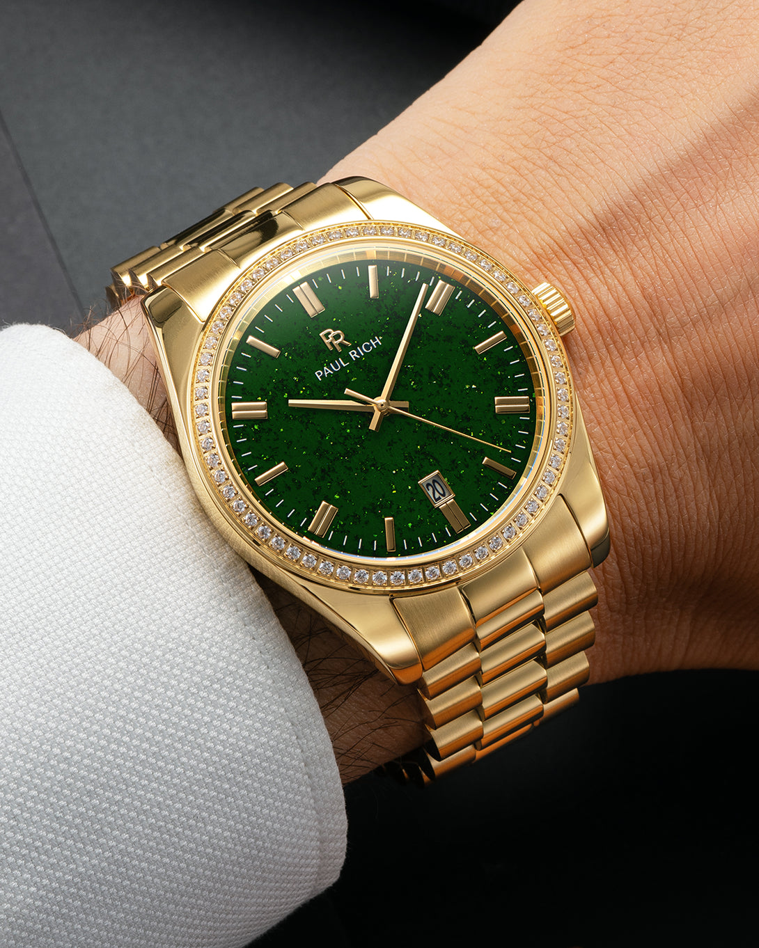 Gold-tone Paul Rich watch with green aventurine dial, diamond-set bezel, and gold link bracelet, worn on wrist with a white textured dress shirt cuff. Gold-tone Paul Rich watch with green aventurine dial, diamond-set bezel, and gold link bracelet, worn on wrist with a white textured dress shirt cuff.