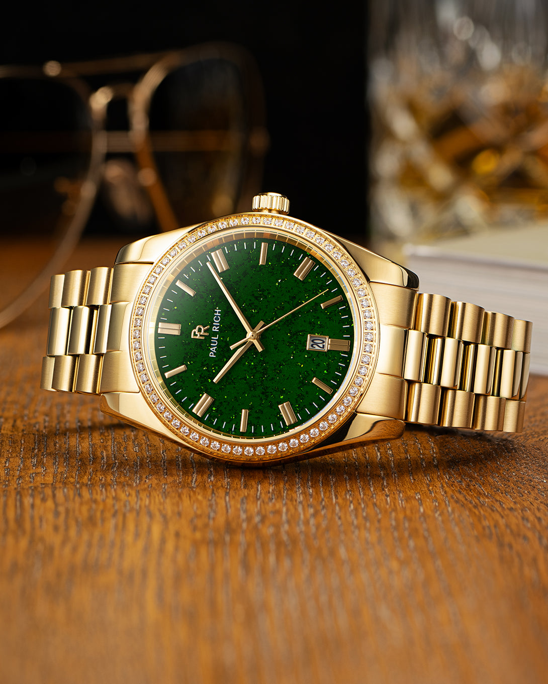 Gold-tone Paul Rich watch with green aventurine dial, diamond-set bezel, and gold link bracelet, displayed on a wooden surface with a pair of sunglasses and a glass tumbler in the background. Gold-tone Paul Rich watch with green aventurine dial, diamond-set bezel, and gold link bracelet, displayed on a wooden surface with a pair of sunglasses and a glass tumbler in the background.