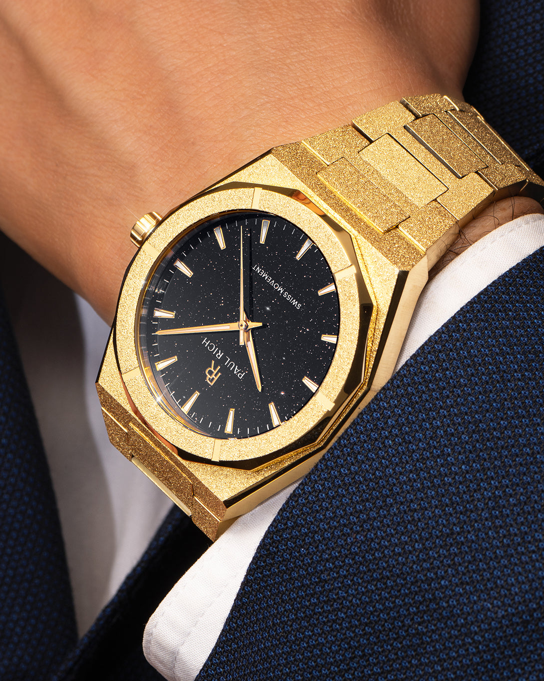 Gold Paul Rich watch with black aventurine dial and frosted finish, worn on wrist with a dark blue textured suit and white cuff. Gold Paul Rich watch with black aventurine dial and frosted finish, worn on wrist with a dark blue textured suit and white cuff.