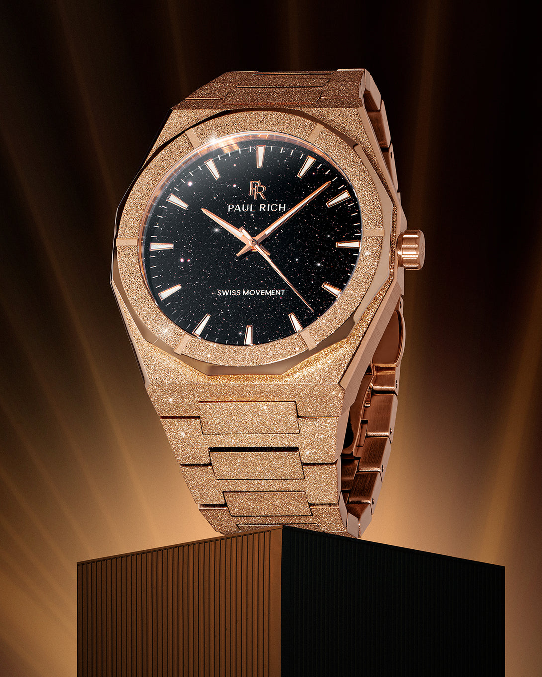 Rose gold Paul Rich watch with fully frosted case and bracelet, black aventurine dial, rose gold markers and hands, and labeled “Swiss Movement,” displayed upright on a pedestal with dramatic golden lighting. Rose gold Paul Rich watch with fully frosted case and bracelet, black aventurine dial, rose gold markers and hands, and labeled “Swiss Movement,” displayed upright on a pedestal with dramatic golden lighting.