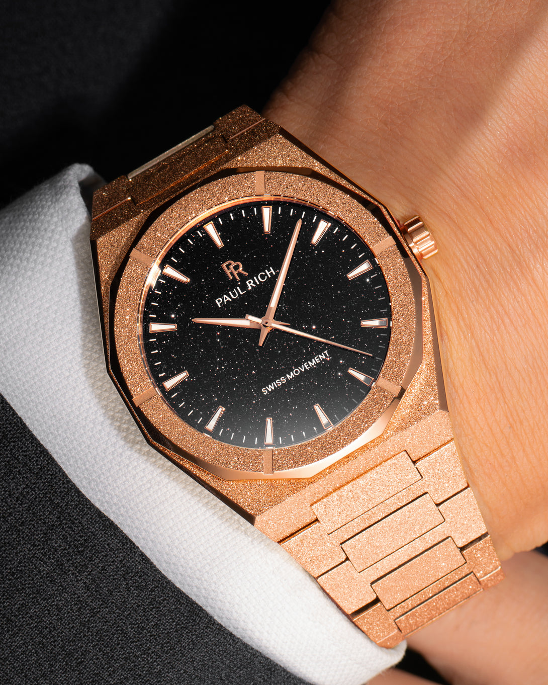 Rose gold Paul Rich watch with fully frosted case and bracelet, blue aventurine dial, rose gold markers and hands, worn on wrist with a white cuff and black suit. Rose gold Paul Rich watch with fully frosted case and bracelet, blue aventurine dial, rose gold markers and hands, worn on wrist with a white cuff and black suit.
