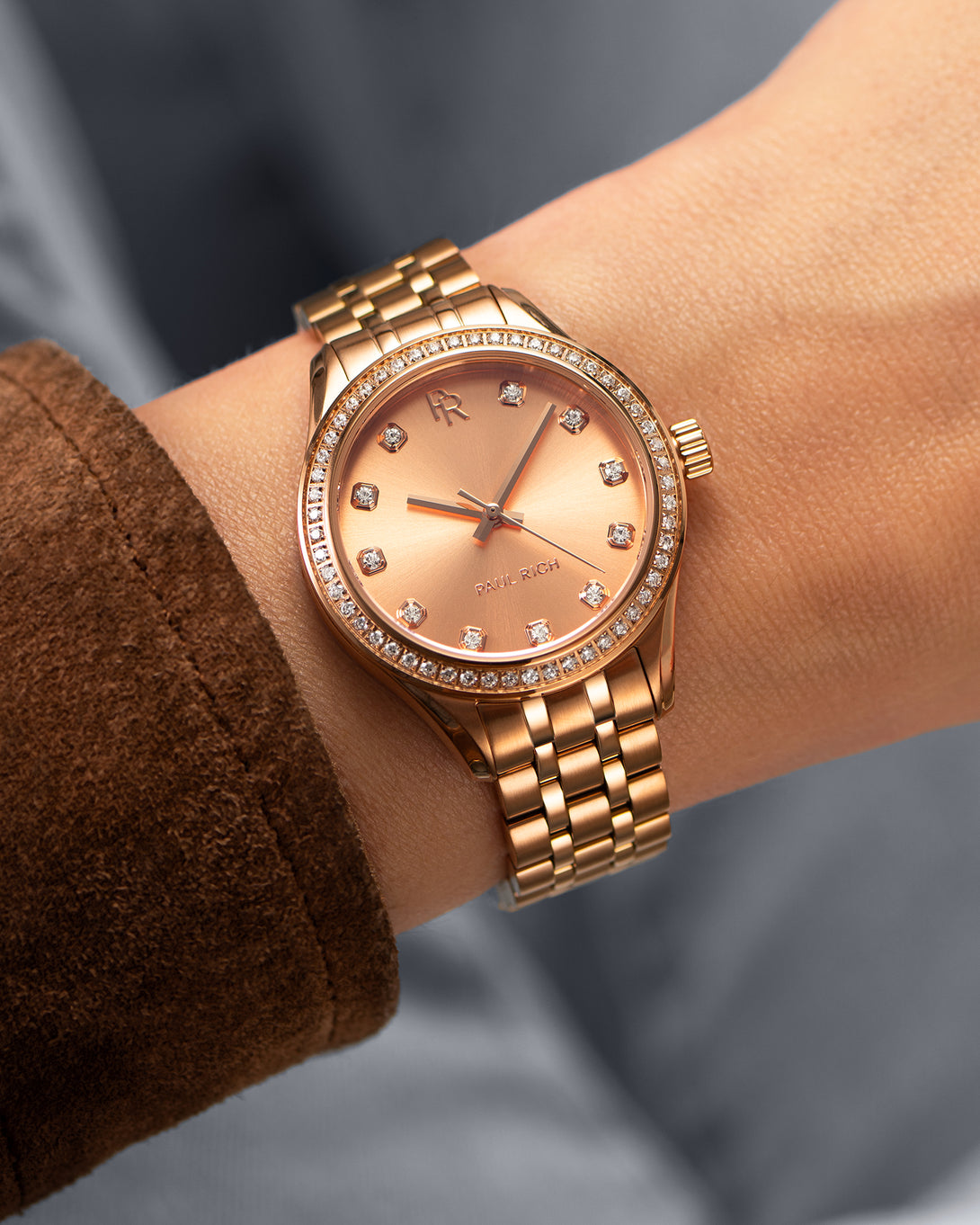 Rose gold Paul Rich watch with a matching sunray dial, diamond hour markers, and gem-set bezel, worn on wrist with a brown suede sleeve. Rose gold Paul Rich watch with a matching sunray dial, diamond hour markers, and gem-set bezel, worn on wrist with a brown suede sleeve.