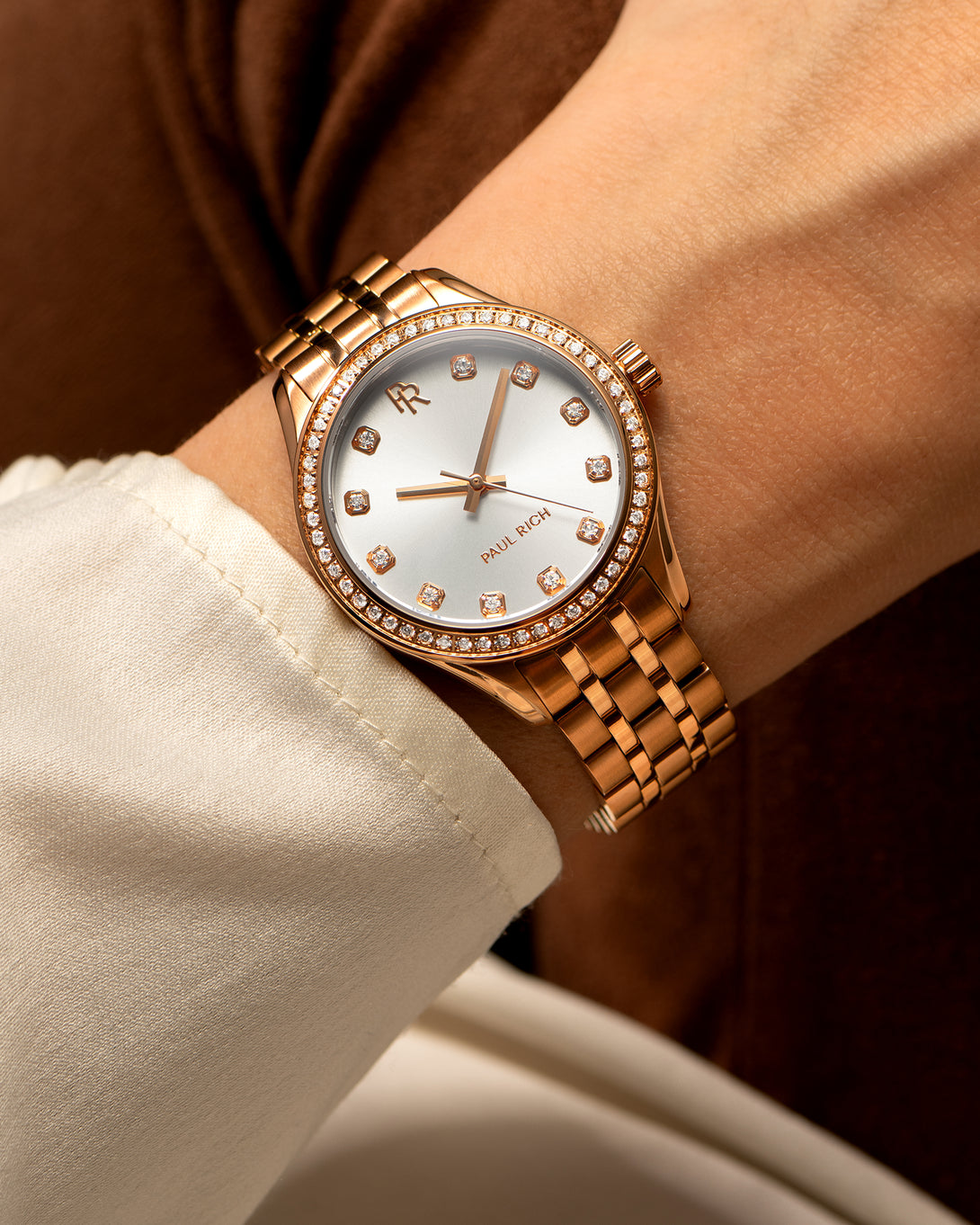 Close-up of a rose gold Paul Rich watch with diamond hour markers and a gem-set bezel, worn on a wrist with a cream-colored sleeve. Close-up of a rose gold Paul Rich watch with diamond hour markers and a gem-set bezel, worn on a wrist with a cream-colored sleeve.