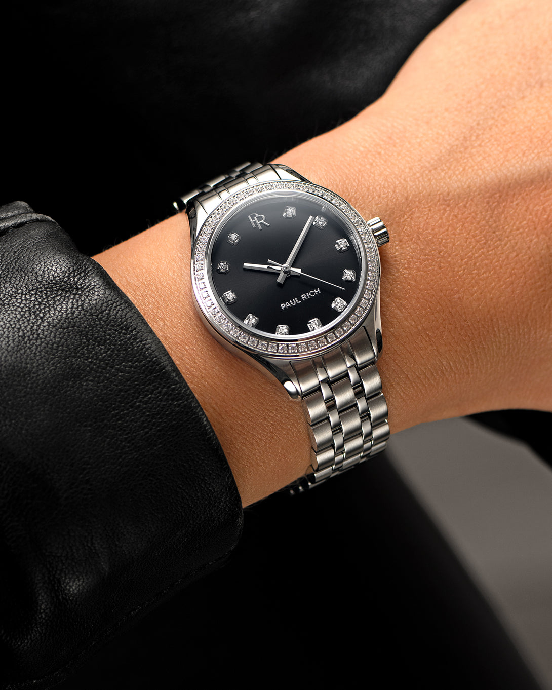 Silver-tone Paul Rich watch with black sunray dial, diamond hour markers, and gem-set bezel, worn on wrist with a black leather sleeve. Silver-tone Paul Rich watch with black sunray dial, diamond hour markers, and gem-set bezel, worn on wrist with a black leather sleeve.