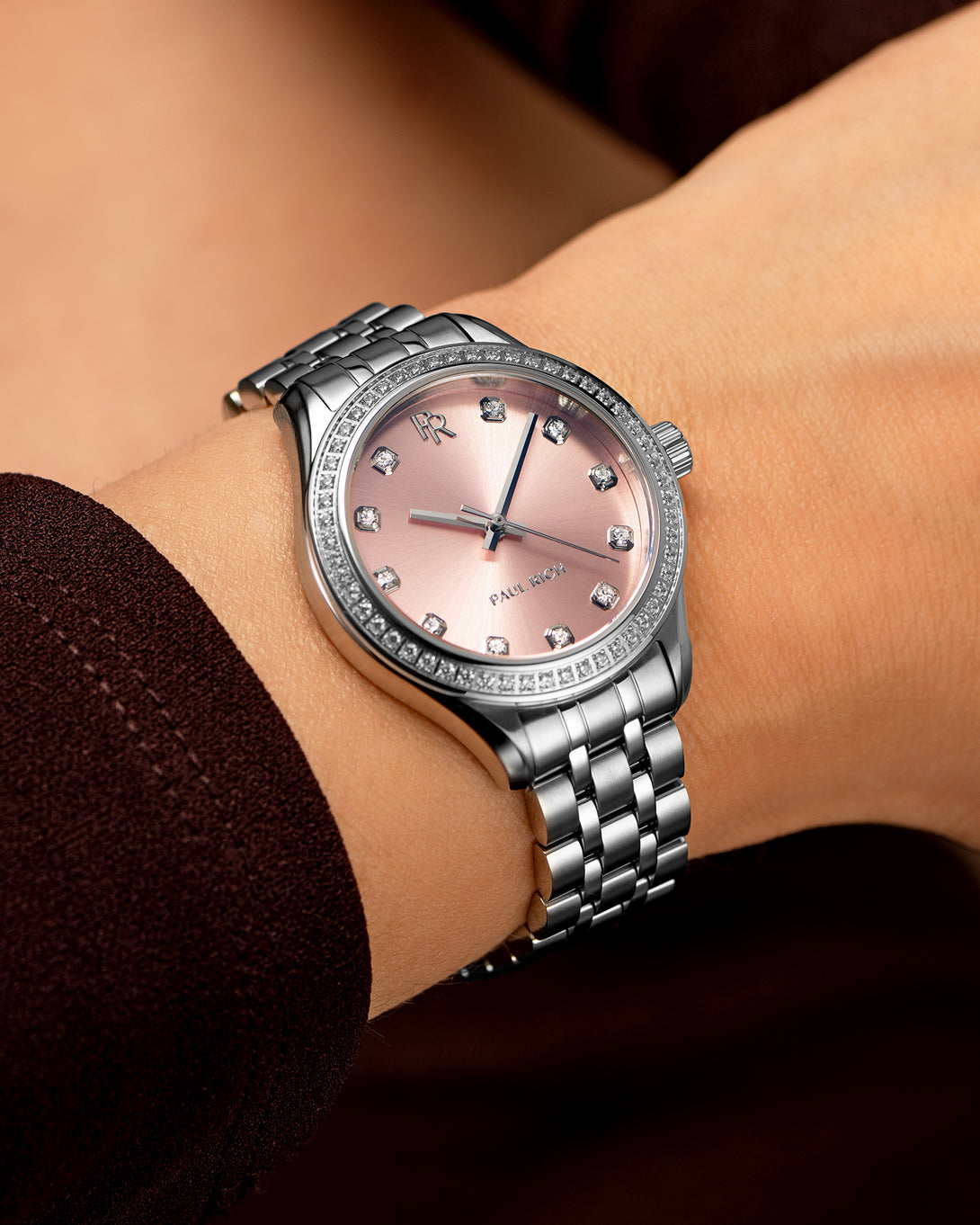Silver-tone Paul Rich watch with pink sunray dial, diamond hour markers, and gem-set bezel, worn on wrist with a dark burgundy sleeve. Silver-tone Paul Rich watch with pink sunray dial, diamond hour markers, and gem-set bezel, worn on wrist with a dark burgundy sleeve.