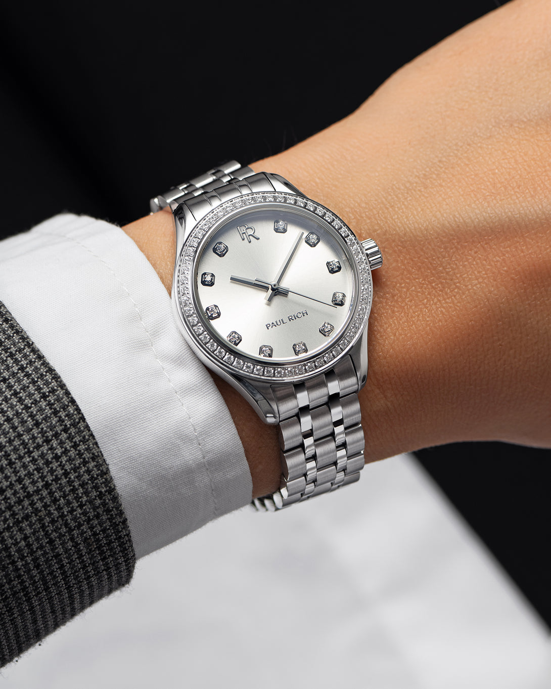 Silver-tone Paul Rich watch with a silver sunray dial, diamond hour markers, and gem-set bezel, worn on wrist with a white shirt and grey check blazer. Silver-tone Paul Rich watch with a silver sunray dial, diamond hour markers, and gem-set bezel, worn on wrist with a white shirt and grey check blazer.
