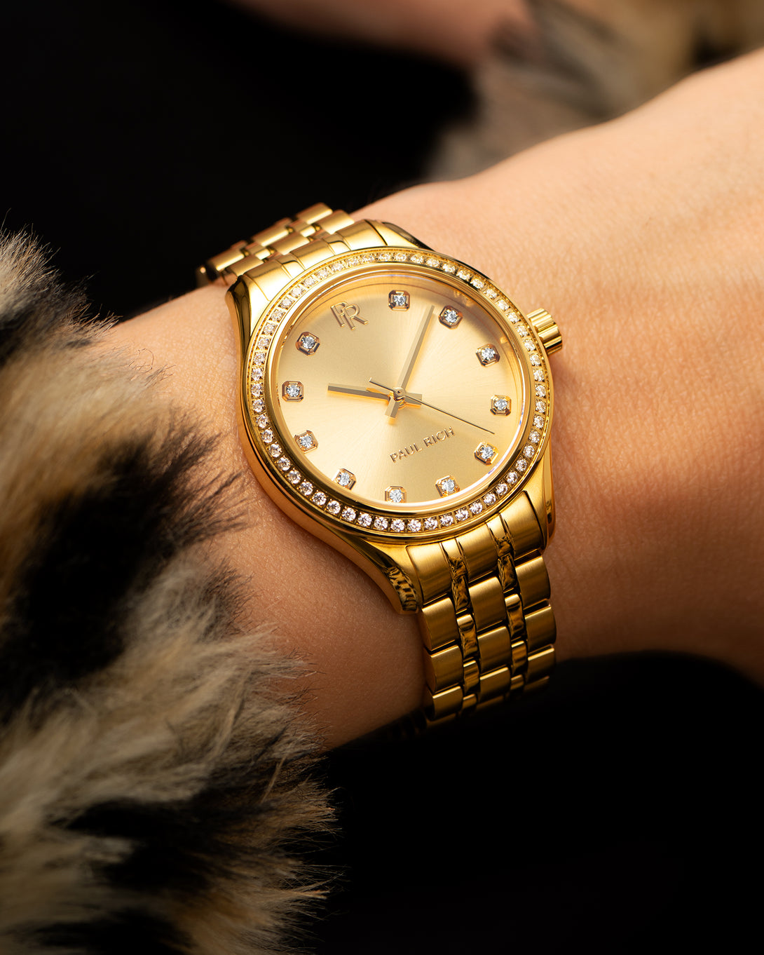 Gold-tone Paul Rich watch with gold sunray dial, diamond hour markers, and gem-set bezel, worn on a wrist with a faux fur coat sleeve. Gold-tone Paul Rich watch with gold sunray dial, diamond hour markers, and gem-set bezel, worn on a wrist with a faux fur coat sleeve.
