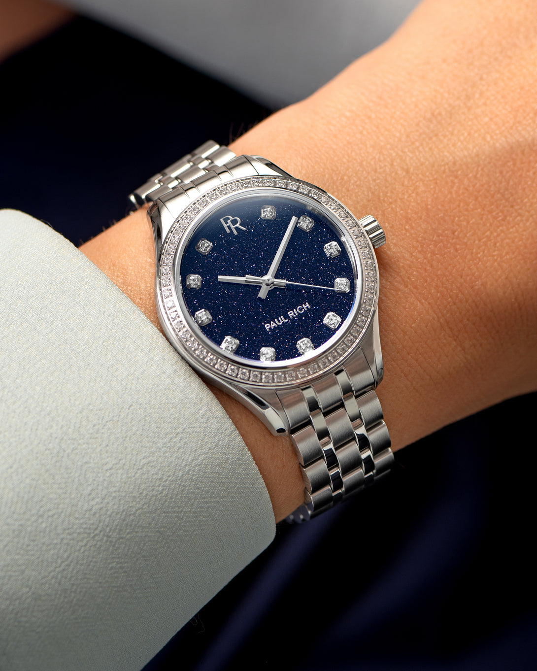 Silver-tone Paul Rich watch with a glittering deep blue aventurine dial, diamond hour markers, and gem-set bezel, worn on wrist with a light blue cuff. Silver-tone Paul Rich watch with a glittering deep blue aventurine dial, diamond hour markers, and gem-set bezel, worn on wrist with a light blue cuff.