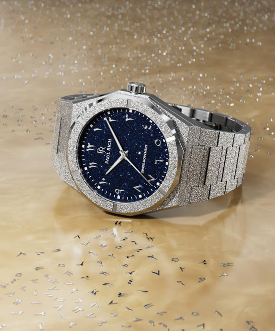 Paul Rich silver frosted watch with blue aventurine dial and Arabic numerals, displayed on gold surface with scattered metal letters. Paul Rich silver frosted watch with blue aventurine dial and Arabic numerals, displayed on gold surface with scattered metal letters.