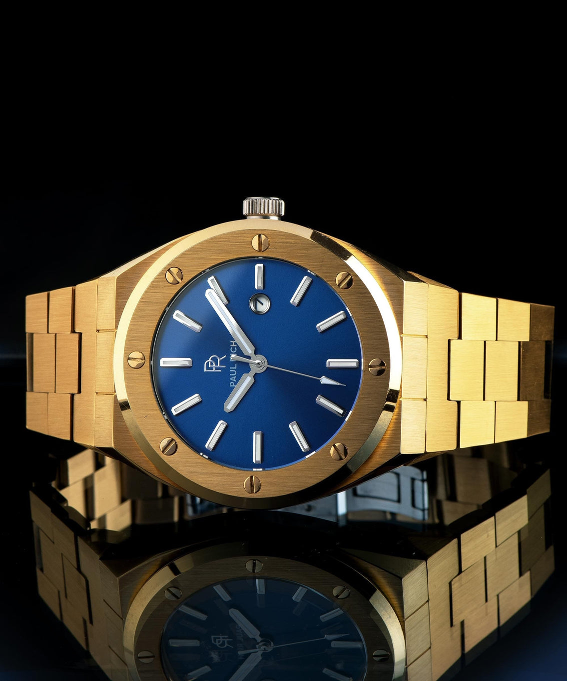 Paul Rich brushed gold watch with matte blue dial and silver markers, displayed horizontally on reflective black surface with crown visible. Paul Rich brushed gold watch with matte blue dial and silver markers, displayed horizontally on reflective black surface with crown visible.