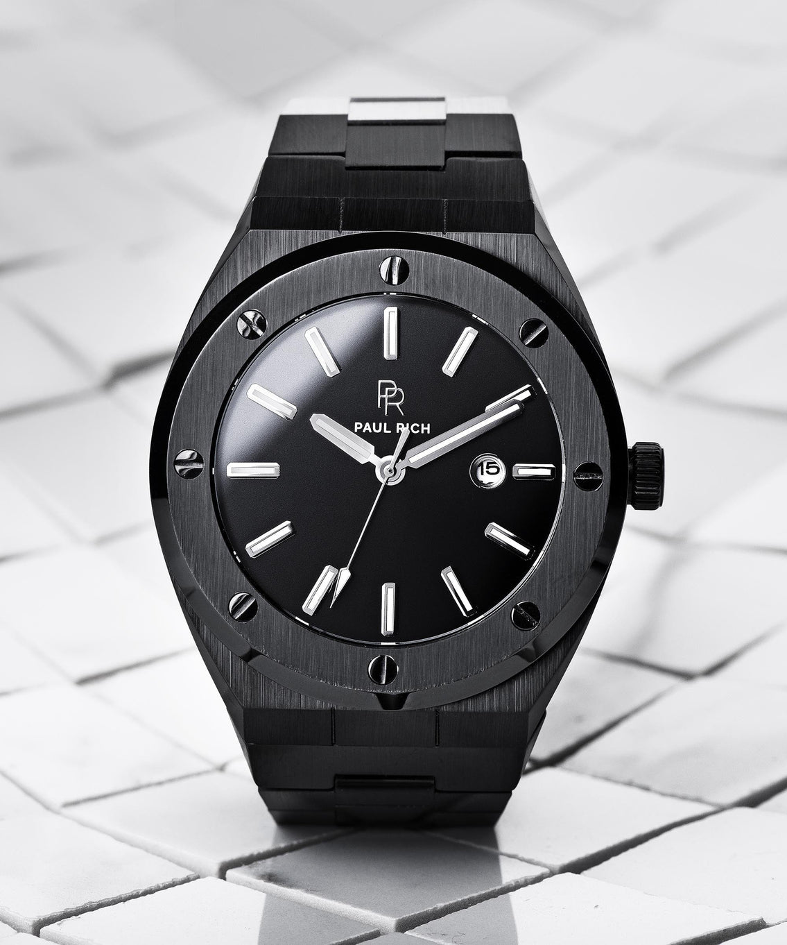 Paul Rich brushed black watch with black sunray dial and date window, displayed upright on white tile surface with soft shadows. Paul Rich brushed black watch with black sunray dial and date window, displayed upright on white tile surface with soft shadows.