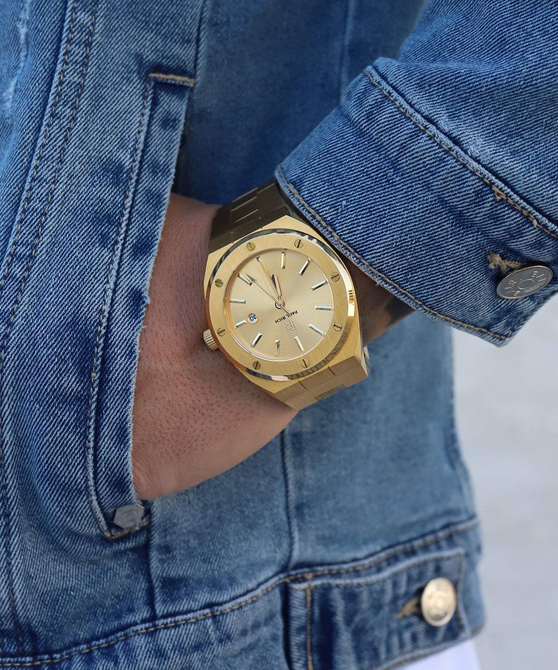 Paul Rich brushed gold watch with gold dial and date window, worn on wrist with blue denim jacket, hand in pocket. Paul Rich brushed gold watch with gold dial and date window, worn on wrist with blue denim jacket, hand in pocket.