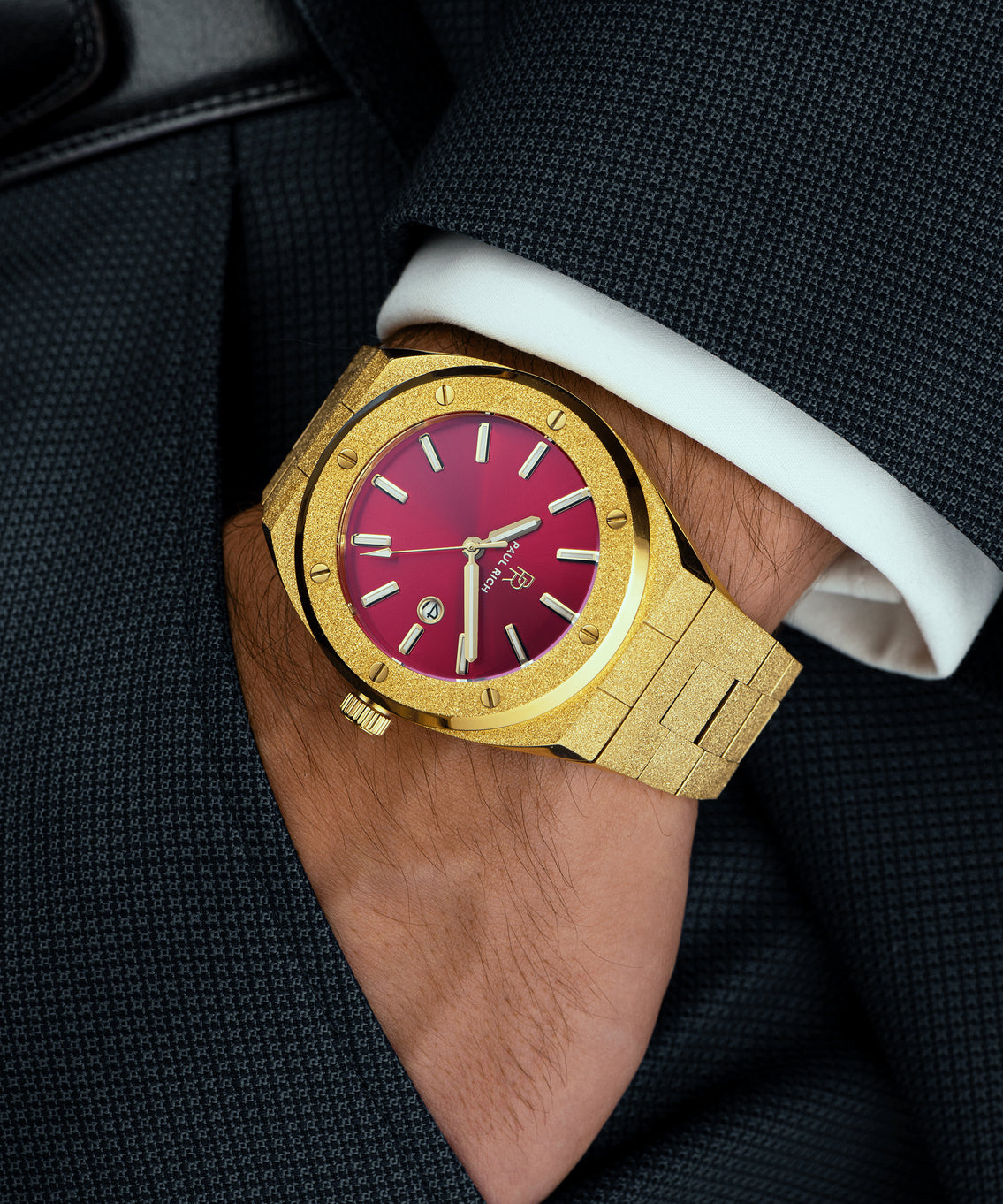 Paul Rich watch with frosted gold case and bracelet, metallic red sunray dial, and date window, worn on wrist with dark textured blazer and white cuff. Paul Rich watch with frosted gold case and bracelet, metallic red sunray dial, and date window, worn on wrist with dark textured blazer and white cuff.