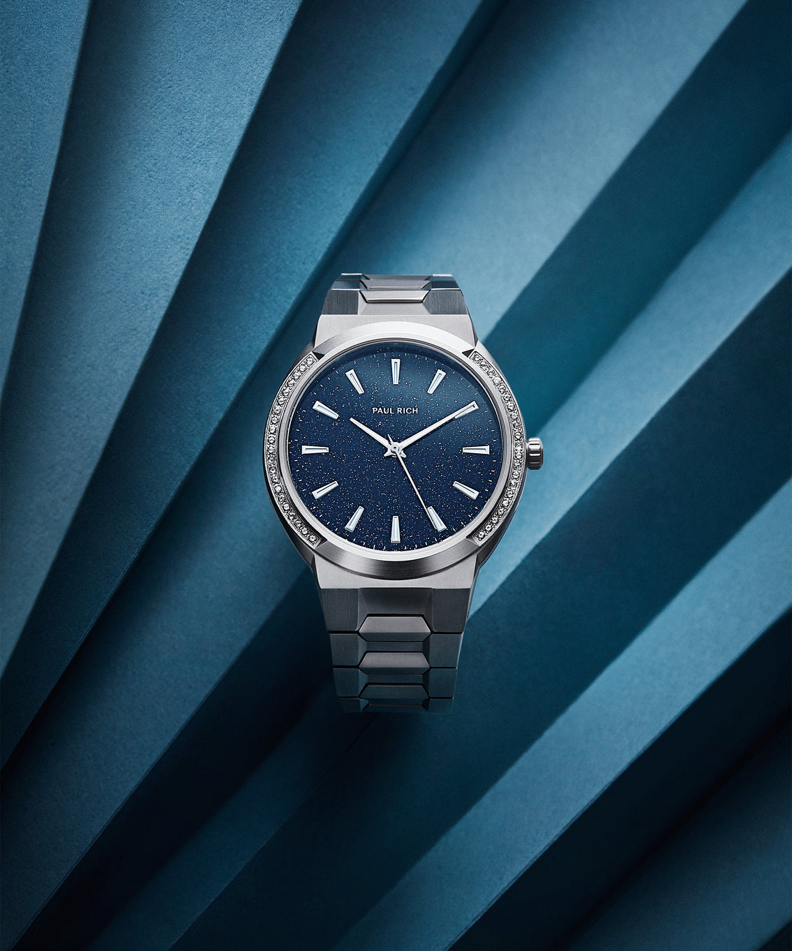 Paul Rich watch with round case, gem-set bezel, blue aventurine dial, and stainless steel bracelet, displayed on folded blue paper background. Paul Rich watch with round case, gem-set bezel, blue aventurine dial, and stainless steel bracelet, displayed on folded blue paper background.