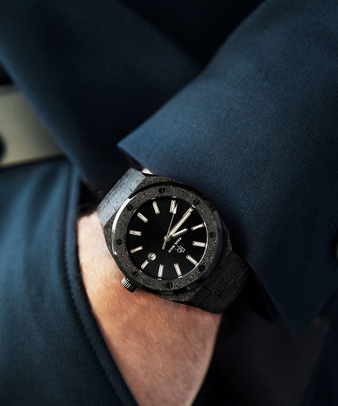 Paul Rich frosted black watch with sunray dial and silver markers, worn on wrist with dark blazer, close-up view. Paul Rich frosted black watch with sunray dial and silver markers, worn on wrist with dark blazer, close-up view.