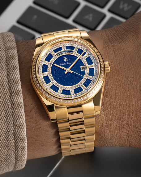 Gold watch with a blue aventurine dial, pavé diamond bezel, and day–date display shown on a wrist with a matching gold bracelet.