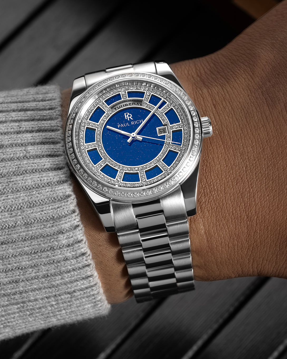 Silver watch with a blue aventurine dial, pavé diamond bezel, and day–date display shown on a wrist with a polished steel bracelet.