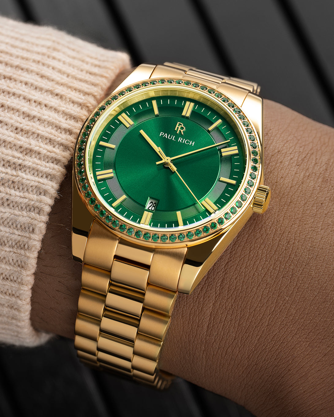 Gold watch with emerald-green dial and green stone bezel on a wrist wearing a beige knit sleeve.