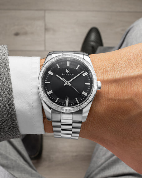 Silver watch with matte black dial, crystal bezel, and jubilee bracelet, worn with a grey suit and black dress shoes.