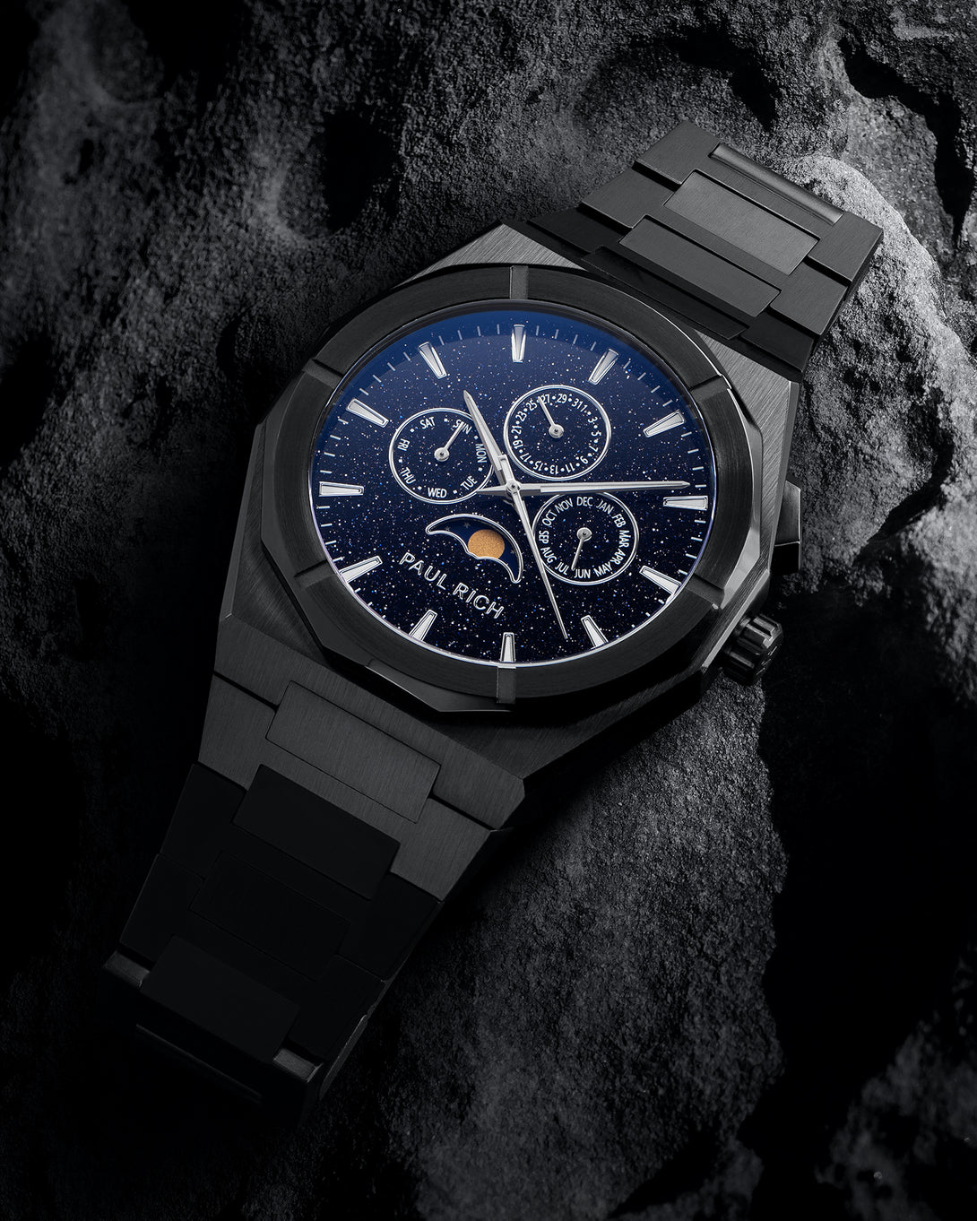 Black steel bracelet watch with speckled blue dial, moonphase at 6 and three calendar subdials; angled on dark textured rock.