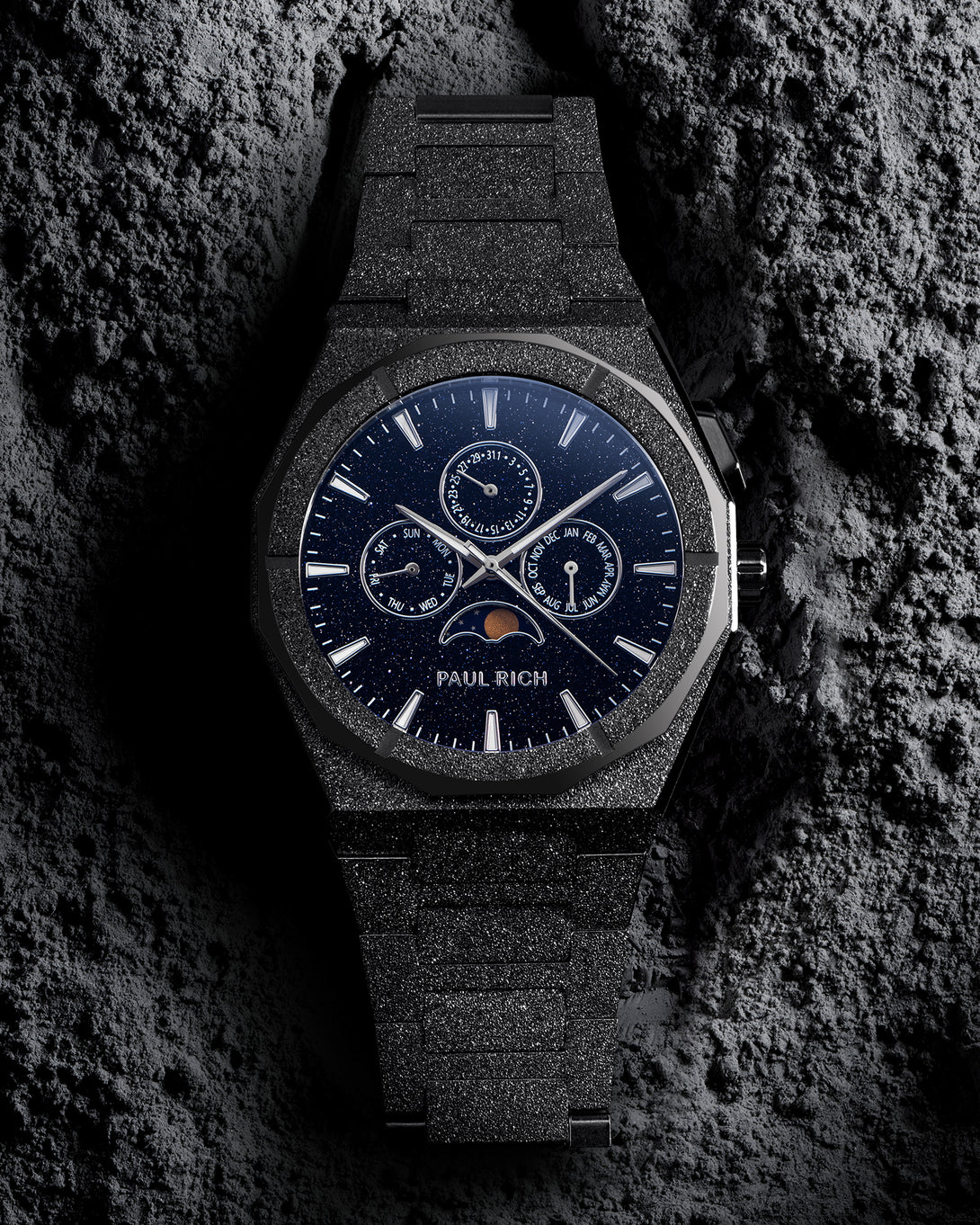 Front view of frosted black bracelet watch with speckled blue dial, moonphase at 6 and three calendar subdials; on gray textured rock.