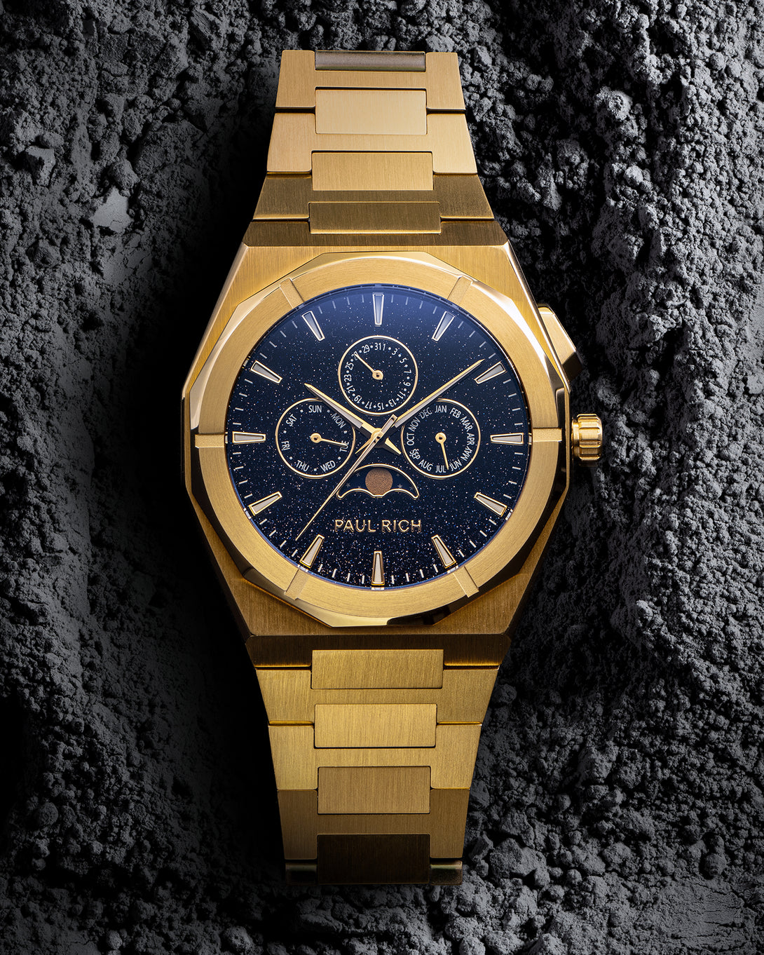 Front view of gold bracelet watch with speckled blue dial, moonphase at 6 and three calendar subdials; brushed links on gray textured rock.