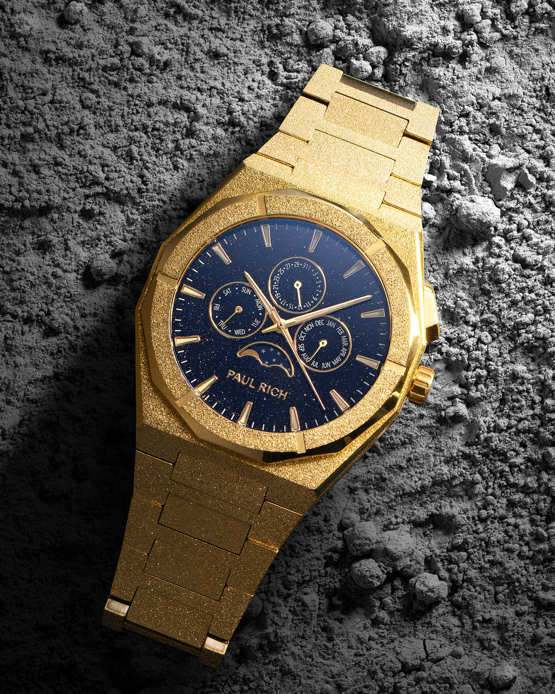 Gold frosted bracelet watch with deep blue speckled dial, moonphase at 6 and three calendar subdials, resting on textured gray rock.