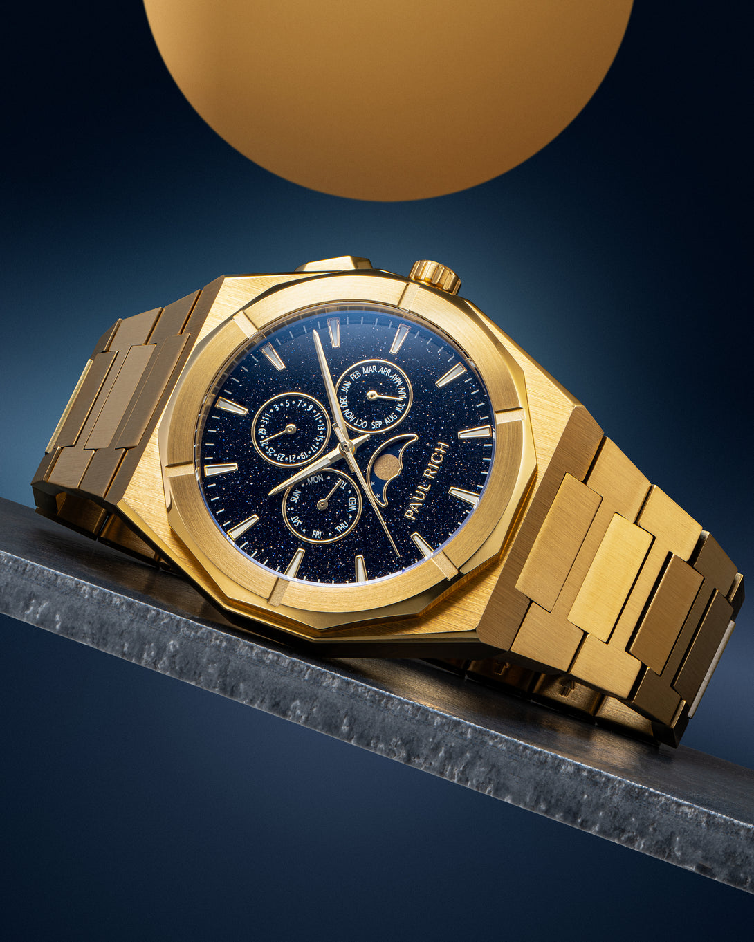 Gold-tone stainless steel watch with blue aventurine dial, moonphase, and subdials, displayed on a metal surface against a dark backdrop.