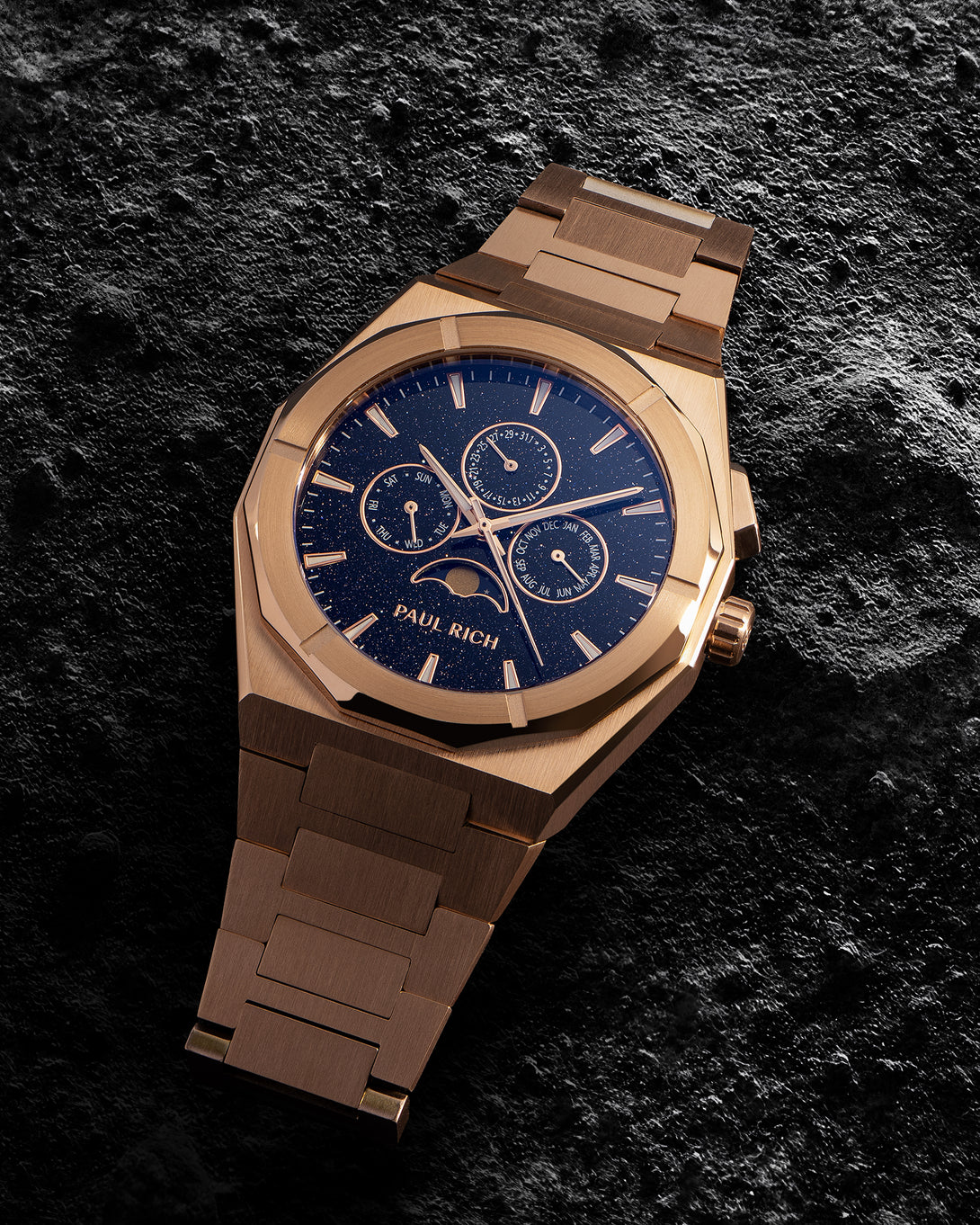 Stainless Steel rose-gold bracelet watch with speckled blue dial, moonphase and calendar subdials; brushed links on dark textured stone.
