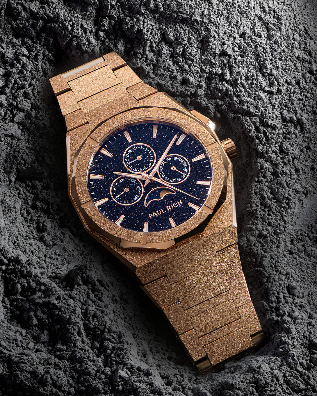 Frosted rose-gold bracelet watch with speckled blue dial, moonphase at 6 and three calendar subdials; angled on gray textured rock.