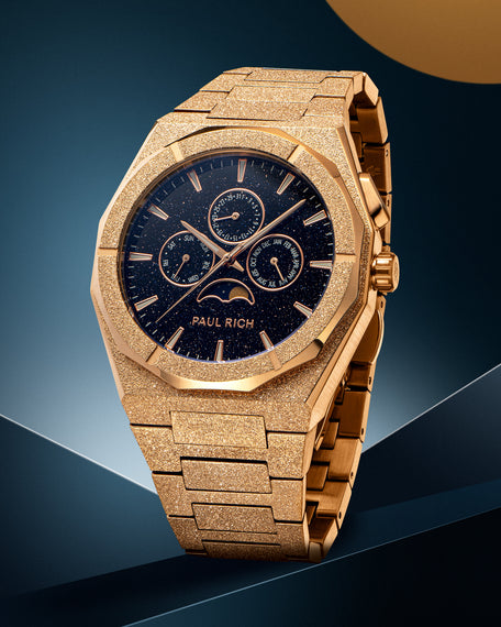 Frosted rose gold-tone stainless steel watch with blue aventurine dial, moonphase, and subdials, displayed on a geometric blue and gold backdrop.