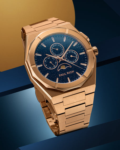 Rose gold-tone stainless steel watch with blue aventurine dial, moonphase, and subdials, displayed on a geometric blue and gold backdrop.
