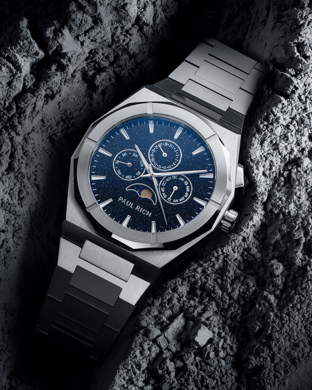 Silver steel bracelet watch with speckled blue dial, moonphase at 6 and three calendar subdials; angled on gray textured rock.