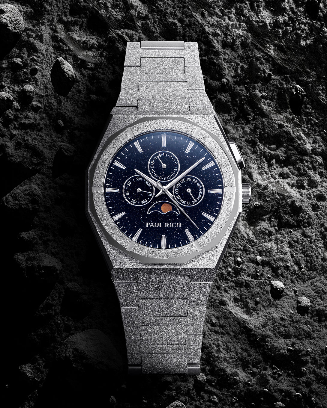 Frosted silver bracelet watch with deep blue speckled dial, moonphase at 6 and three calendar subdials, set on dark textured rock.