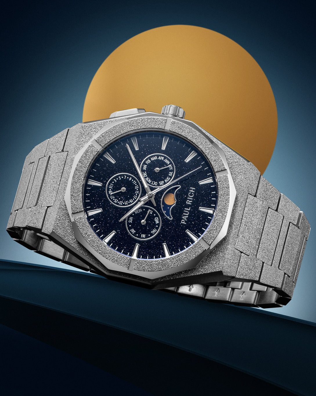 Frosted silver stainless steel watch with blue aventurine dial, moonphase, and subdials, displayed against a blue backdrop with a gold circle.