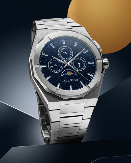 Silver stainless steel watch with blue aventurine dial, moonphase, and subdials, displayed on a geometric silver and blue backdrop with a gold circle.