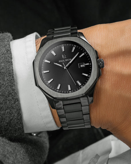Black Paul Rich watch with black sunray dial, steel bracelet, and date window shown on wrist in white shirt and grey coat.