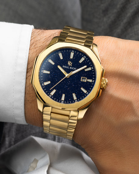 Gold Paul Rich watch with blue aventurine dial, gold bracelet, and date window, worn on wrist with white shirt and grey trousers.