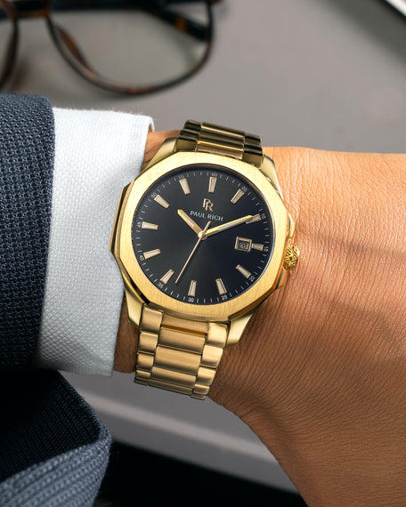 Gold Paul Rich watch with black sunray dial, gold bracelet, and date window, worn on wrist with white cuff and navy textured suit.