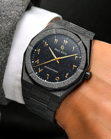 Close-up of Paul Rich frosted black watch with gold Arabic numerals and black aventurine dial, worn with white shirt and grey coat.