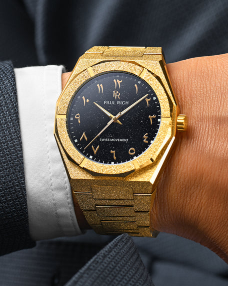 Close-up of Paul Rich frosted gold watch with Arabic numerals and black aventurine dial, worn on wrist with dark textured suit.