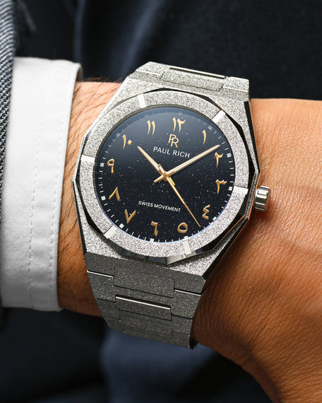 Close-up of Paul Rich frosted silver watch with gold Arabic numerals and black aventurine dial, worn with a white cuff and dark jacket.