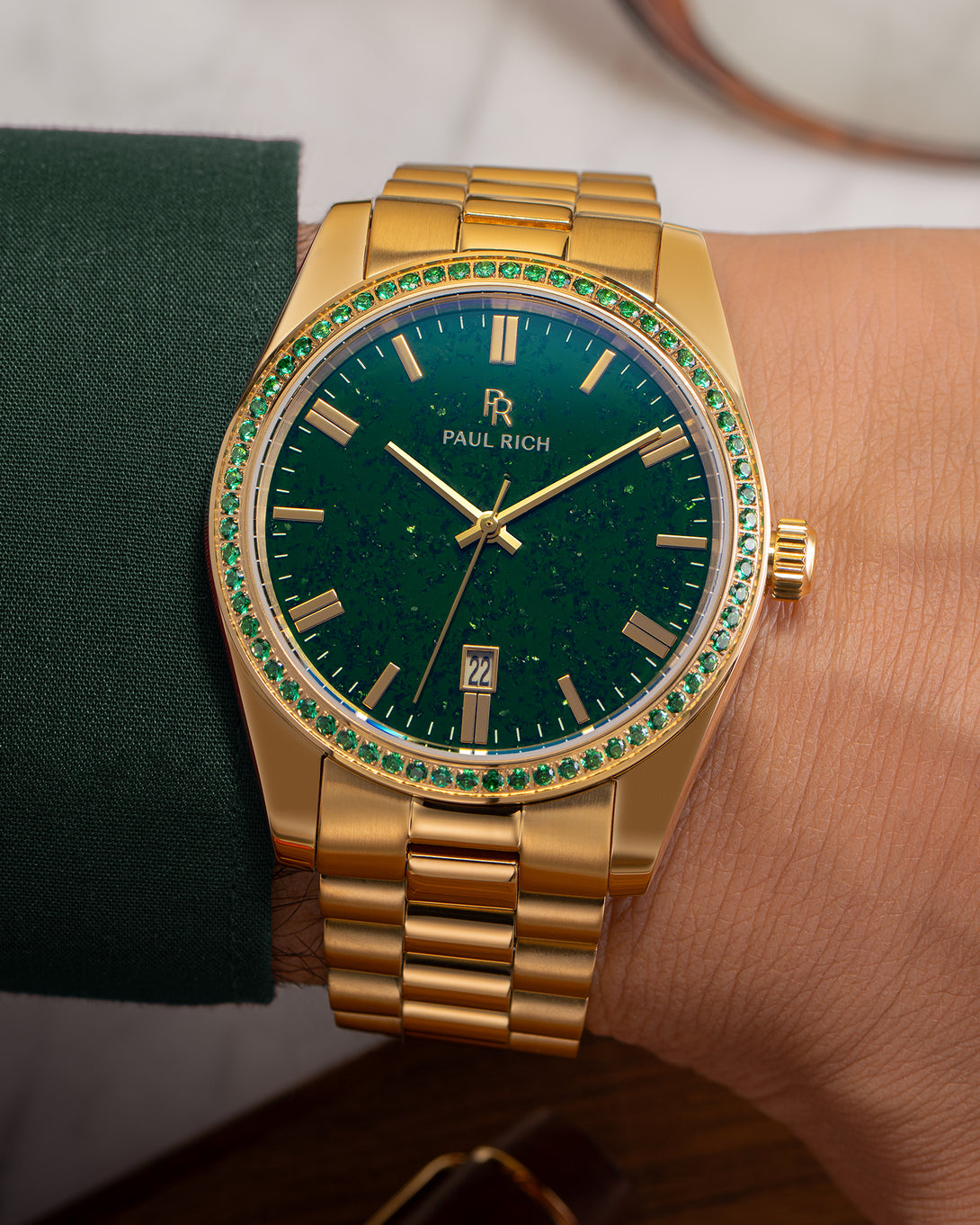 Gold watch with green aventurine dial and green gemstone-set bezel on a wrist wearing a dark green sleeve, showing the Paul Rich logo and date window.