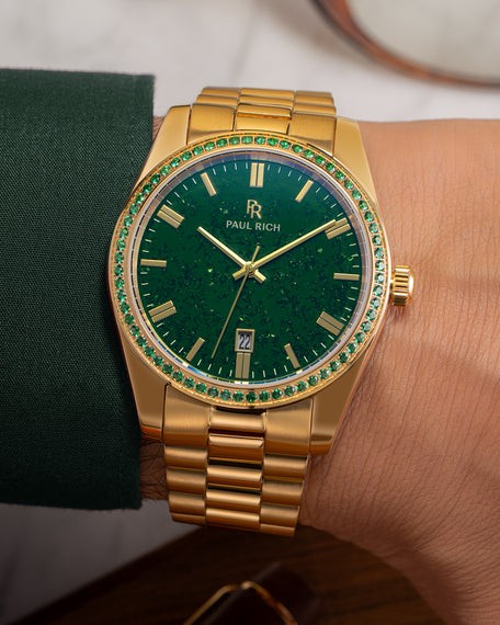 Gold watch with green aventurine dial and green gemstone-set bezel on a wrist wearing a dark green sleeve, showing the Paul Rich logo and date window.