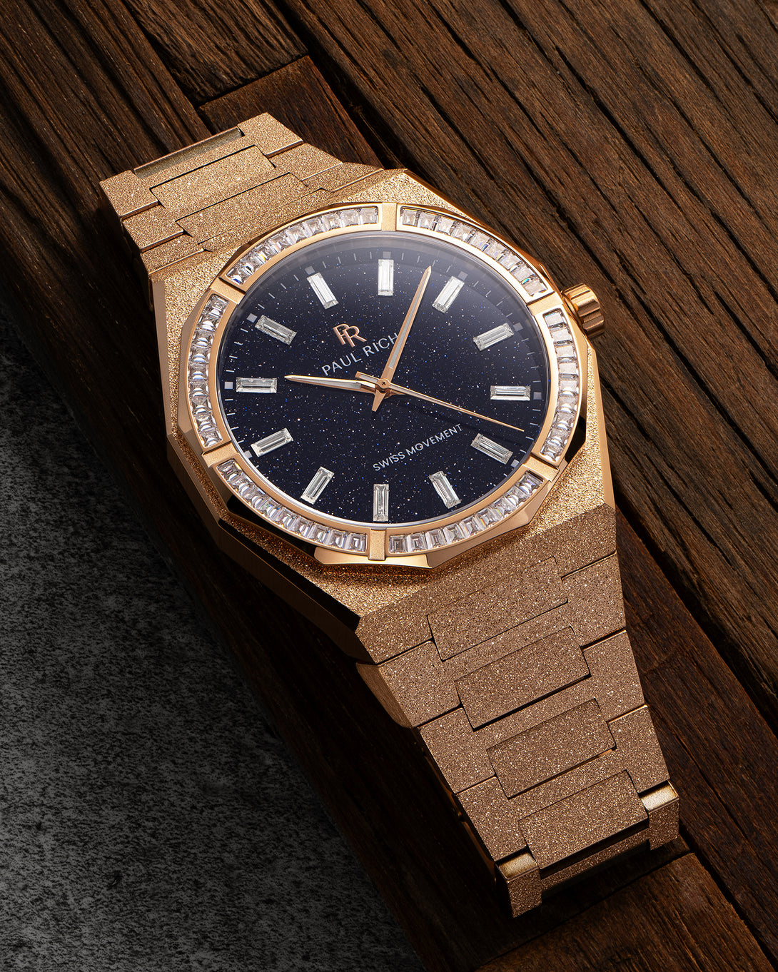 Rose gold frosted watch with aventurine dial and baguette-cut moissanite bezel, displayed on a dark wooden surface.