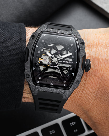 Black frosted tonneau skeleton watch with black strap, shown on wrist beside a black sleeve and keyboard.