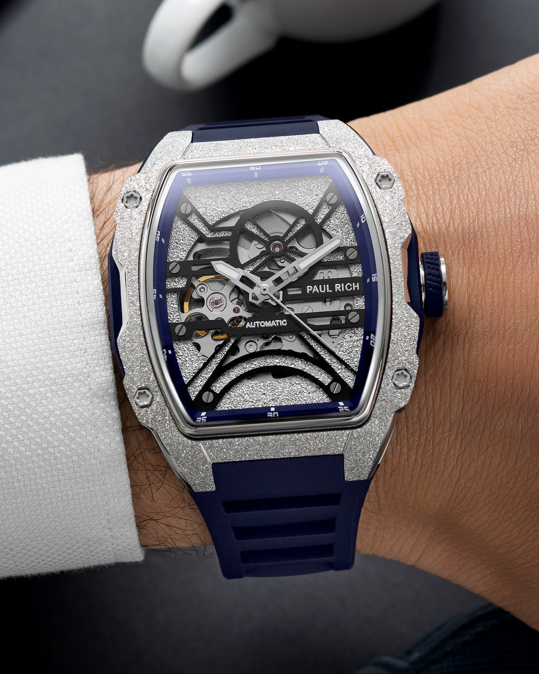 Silver frosted tonneau skeleton watch with blue strap, worn on wrist beside a white textured shirt cuff and coffee cup.