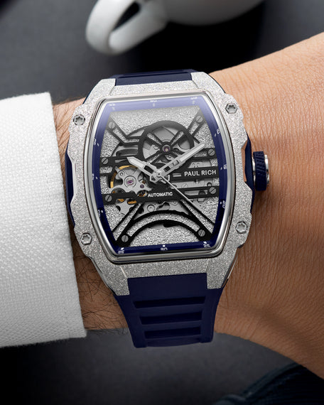 Silver frosted tonneau skeleton watch with blue strap, worn on wrist beside a white textured shirt cuff and coffee cup.